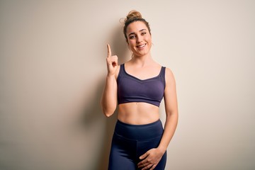 Young beautiful blonde sportswoman doing sport wearing sportswear over white background showing and pointing up with finger number one while smiling confident and happy.