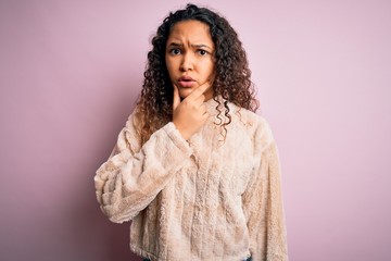 Young beautiful woman with curly hair wearing casual sweater standing over pink background Looking fascinated with disbelief, surprise and amazed expression with hands on chin