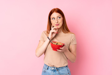 Young redhead woman over isolated pink background holding a bowl of cereals and thinking