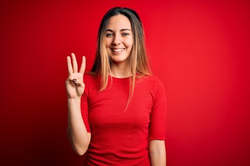 Young beautiful blonde woman with blue eyes wearing casual t-shirt over red background showing and pointing up with fingers number three while smiling confident and happy.