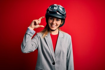 Young beautiful blonde motorcyclist woman wearing motorcycle helmet over red background smiling and confident gesturing with hand doing small size sign with fingers looking and the camera. Measure 