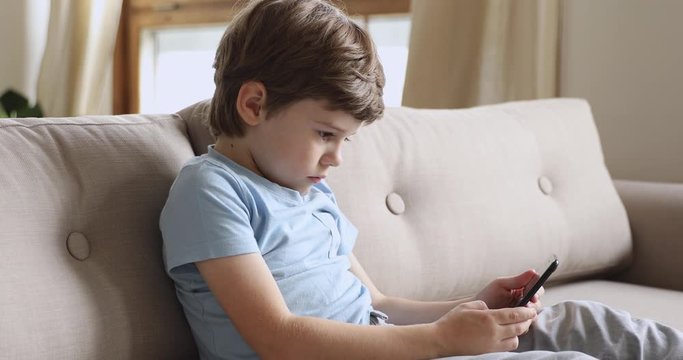 Happy small kid boy sitting on couch, using funny photo editing mobile applications. Smiling little child playing online games on smartphone, spending free time in social networks alone at home.