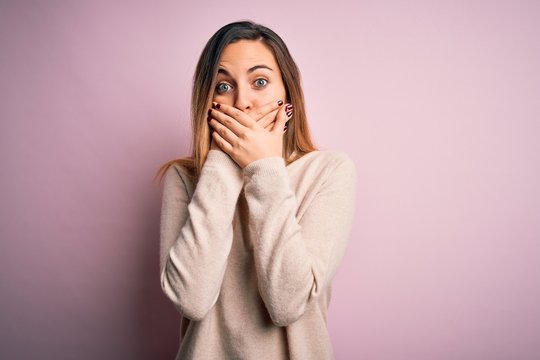 Beautiful blonde woman with blue eyes wearing turtleneck sweater over pink background shocked covering mouth with hands for mistake. Secret concept.