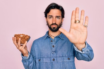 Young handsome hispanic man with bear holding natural ingredient healthy almonds with open hand doing stop sign with serious and confident expression, defense gesture