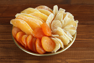 Slices of candied ginger root, pieces of dried mango fruits and dried apricots lying on a golden plate on a wooden table. Closeup. Macro