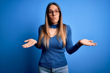 Young beautiful blonde woman with blue eyes wearing glasses standing over blue background clueless...