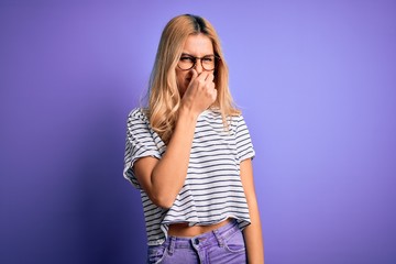 Young beautiful blonde woman wearing striped t-shirt and glasses over purple background smelling something stinky and disgusting, intolerable smell, holding breath with fingers on nose. Bad smell