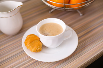 White mug with freshly brewed coffee and a croissant in the kitchen. Healthy breakfast.