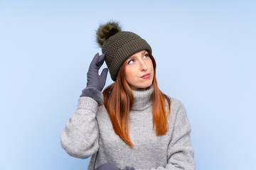 Young redhead Russian woman with winter hat over isolated blue background having doubts and with confuse face expression