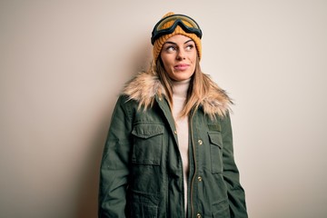 Young brunette skier woman wearing snow clothes and ski goggles over white background smiling looking to the side and staring away thinking.