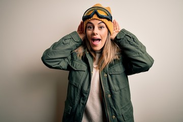Young brunette skier woman wearing snow clothes and ski goggles over white background Smiling pulling ears with fingers, funny gesture. Audition problem
