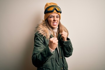 Young brunette skier woman wearing snow clothes and ski goggles over white background Ready to fight with fist defense gesture, angry and upset face, afraid of problem