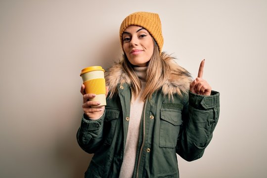Young beautiful brunette woman wearing snow clothes drinking cup of coffee surprised with an idea or question pointing finger with happy face, number one