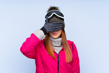 Skier redhead woman with snowboarding glasses over isolated blue wall covering eyes by hands