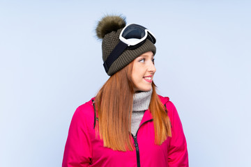 Skier redhead woman with snowboarding glasses over isolated blue wall looking to the side
