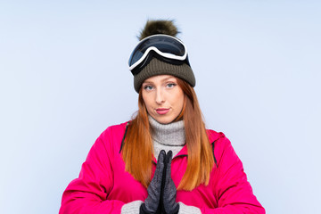 Skier redhead woman with snowboarding glasses over isolated blue wall pleading