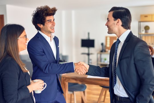 Young beautiful group of business workers smiling happy and confident. Standing with smile on face drinking coffee and shaking hands for agreement at the office