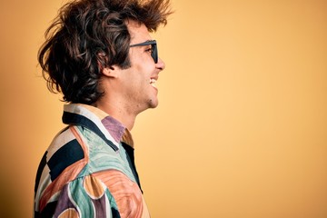 Young man on vacation wearing colorful shirt and glasses over isolated yellow background looking to side, relax profile pose with natural face with confident smile.