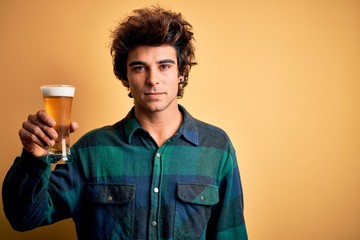 Young handsome man drinking glass of beer standing over isolated yellow background with a confident expression on smart face thinking serious