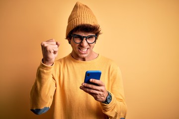 Fototapeta premium Young handsome man using smartphone wearing glasses over isolated yellow background annoyed and frustrated shouting with anger, crazy and yelling with raised hand, anger concept
