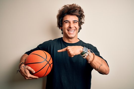 Young handsome sportsman holding basketball ball standing over isolated white background very happy pointing with hand and finger