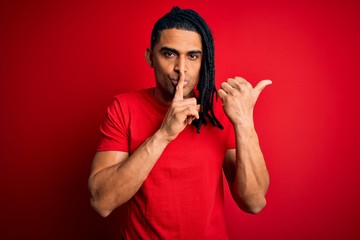 Young handsome african american afro man with dreadlocks wearing red casual t-shirt asking to be quiet with finger on lips pointing with hand to the side. Silence and secret concept.