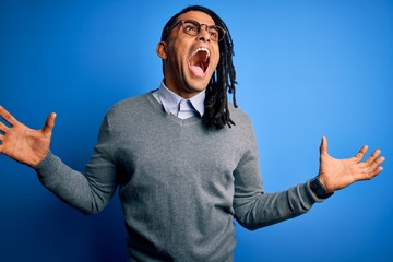 Fototapeta premium Young handsome african american man with dreadlocks wearing casual sweater and glasses crazy and mad shouting and yelling with aggressive expression and arms raised. Frustration concept.