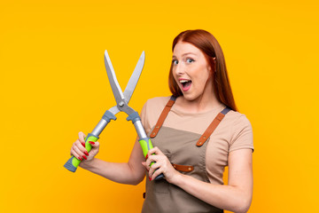 Young redhead gardener woman withpruning shears over isolated yellow background