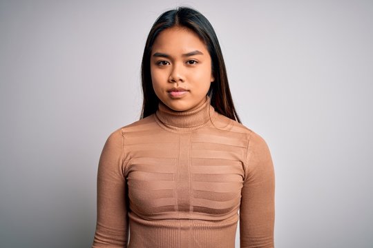 Young beautiful asian girl wearing casual turtleneck sweater standing over white background with serious expression on face. Simple and natural looking at the camera.