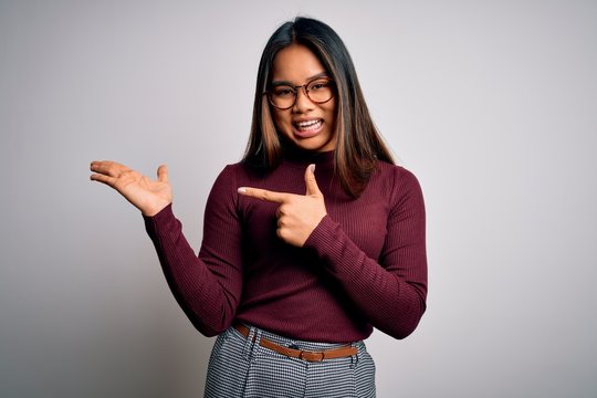 Beautiful asian business woman wearing casual sweater and glasses over white background amazed and smiling to the camera while presenting with hand and pointing with finger.