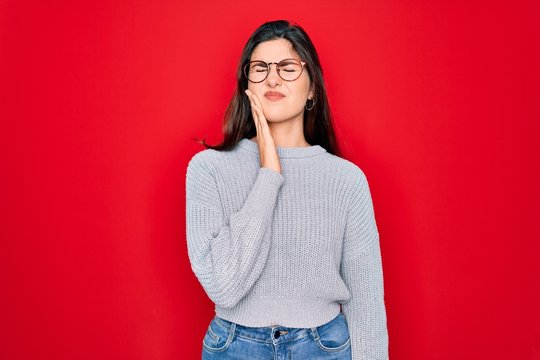 Young beautiful brunette woman wearing casual sweater over red background touching mouth with hand with painful expression because of toothache or dental illness on teeth. Dentist