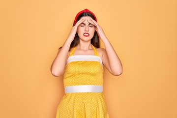 Young beautiful pin up woman wearing 50s fashion vintage dress over yellow background with hand on head for pain in head because stress. Suffering migraine.