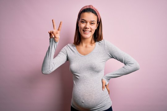 Young beautiful teenager girl pregnant expecting baby over isolated pink background smiling looking to the camera showing fingers doing victory sign. Number two.