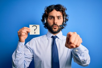 Young call center agent man with beard working using headset holding question mark reminder pointing with finger to the camera and to you, hand sign, positive and confident gesture from the front