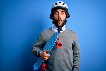 Handsome skater man with beard wearing security helmet holding skate over blue background scared in shock with a surprise face, afraid and excited with fear expression