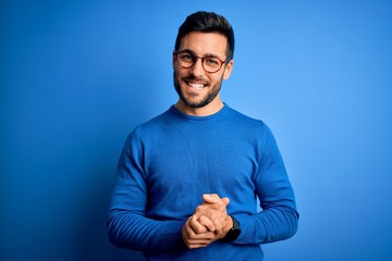 Young handsome man with beard wearing casual sweater and glasses over blue background with hands...