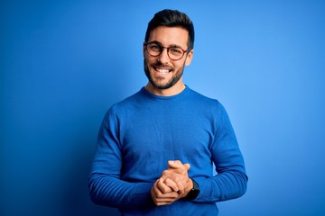 Obraz premium Young handsome man with beard wearing casual sweater and glasses over blue background with hands together and crossed fingers smiling relaxed and cheerful. Success and optimistic