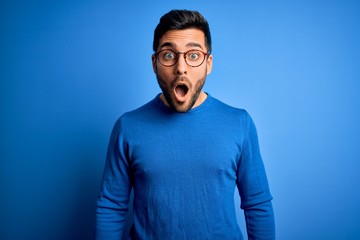 Young handsome man with beard wearing casual sweater and glasses over blue background afraid and...