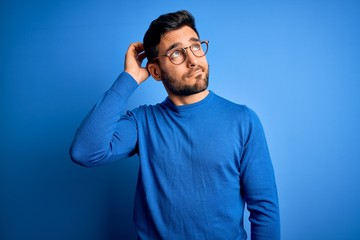 Young handsome man with beard wearing casual sweater and glasses over blue background confuse and...