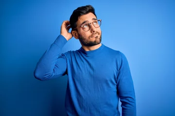 Fototapeten Young handsome man with beard wearing casual sweater and glasses over blue background confuse and wondering about question. Uncertain with doubt, thinking with hand on head. Pensive concept. © Krakenimages.com