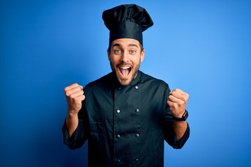Young handsome chef man with beard wearing cooker uniform and hat over blue background celebrating surprised and amazed for success with arms raised and open eyes. Winner concept.