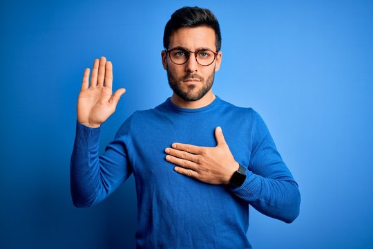 Young handsome man with beard wearing casual sweater and glasses over blue background Swearing with hand on chest and open palm, making a loyalty promise oath