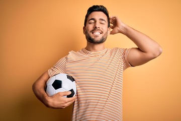 Handsome player man with beard playing soccer holding footballl ball over yellow background smiling confident touching hair with hand up gesture, posing attractive and fashionable