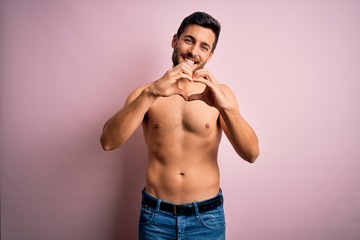 Young handsome strong man with beard shirtless standing over isolated pink background smiling in love doing heart symbol shape with hands. Romantic concept.