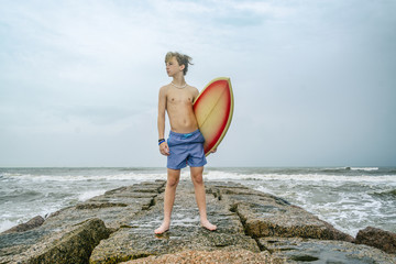 Young teen surfer holding surfboard on rocky coast