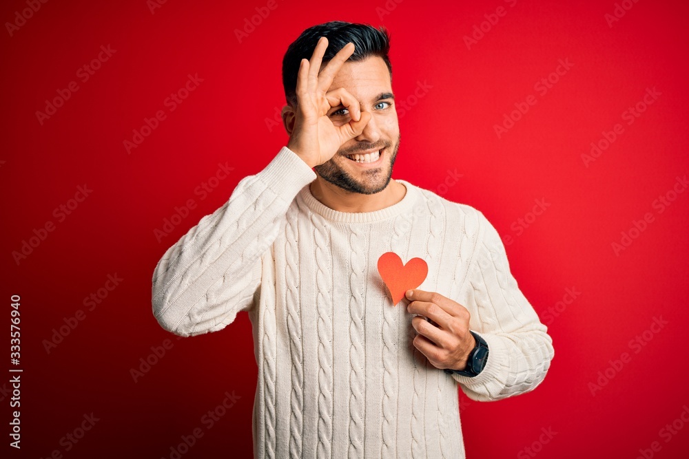 Canvas Prints Young handsome man holding red shape heart as romantic and health symbol with happy face smiling doing ok sign with hand on eye looking through fingers