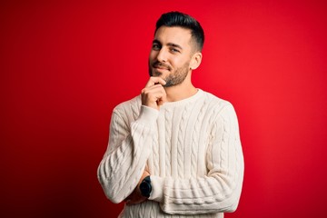 Young handsome man wearing casual white sweater standing over isolated red background looking...
