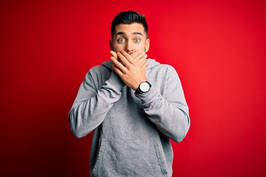 Young handsome sportsman wearing sweatshirt standing over isolated red background shocked covering mouth with hands for mistake. Secret concept.