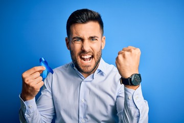 Young handsome man holding blue ribbon as prostate campaing support over blue background annoyed...