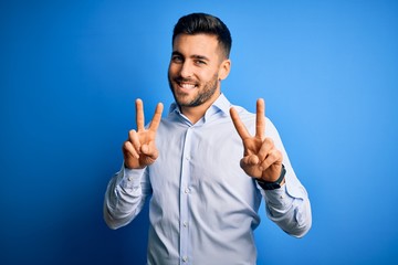 Young handsome man wearing elegant shirt standing over isolated blue background smiling looking to the camera showing fingers doing victory sign. Number two.